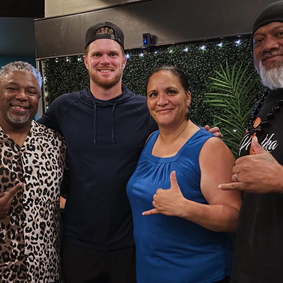 Four smiling people posing together, two making shaka signs, against a green leafy wall.