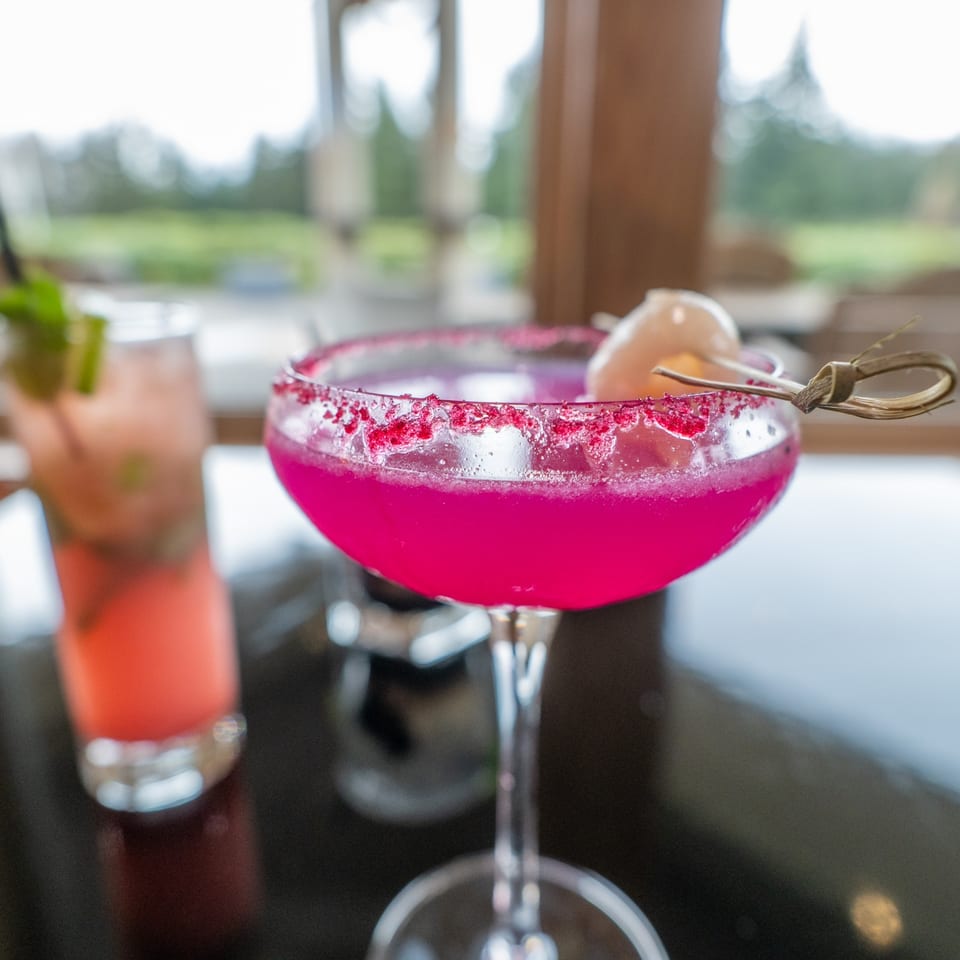 A vibrant pink cocktail with a sugared rim and lychee garnish, next to another drink.