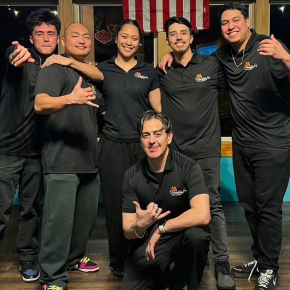 Six restaurant staff members in black polo shirts with a logo, smiling and posing together.