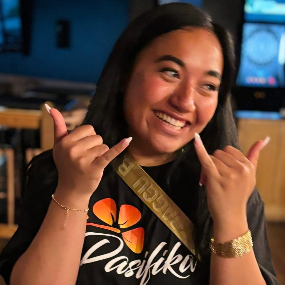 Smiling woman in a black Pasifika shirt making shaka signs with both hands.
