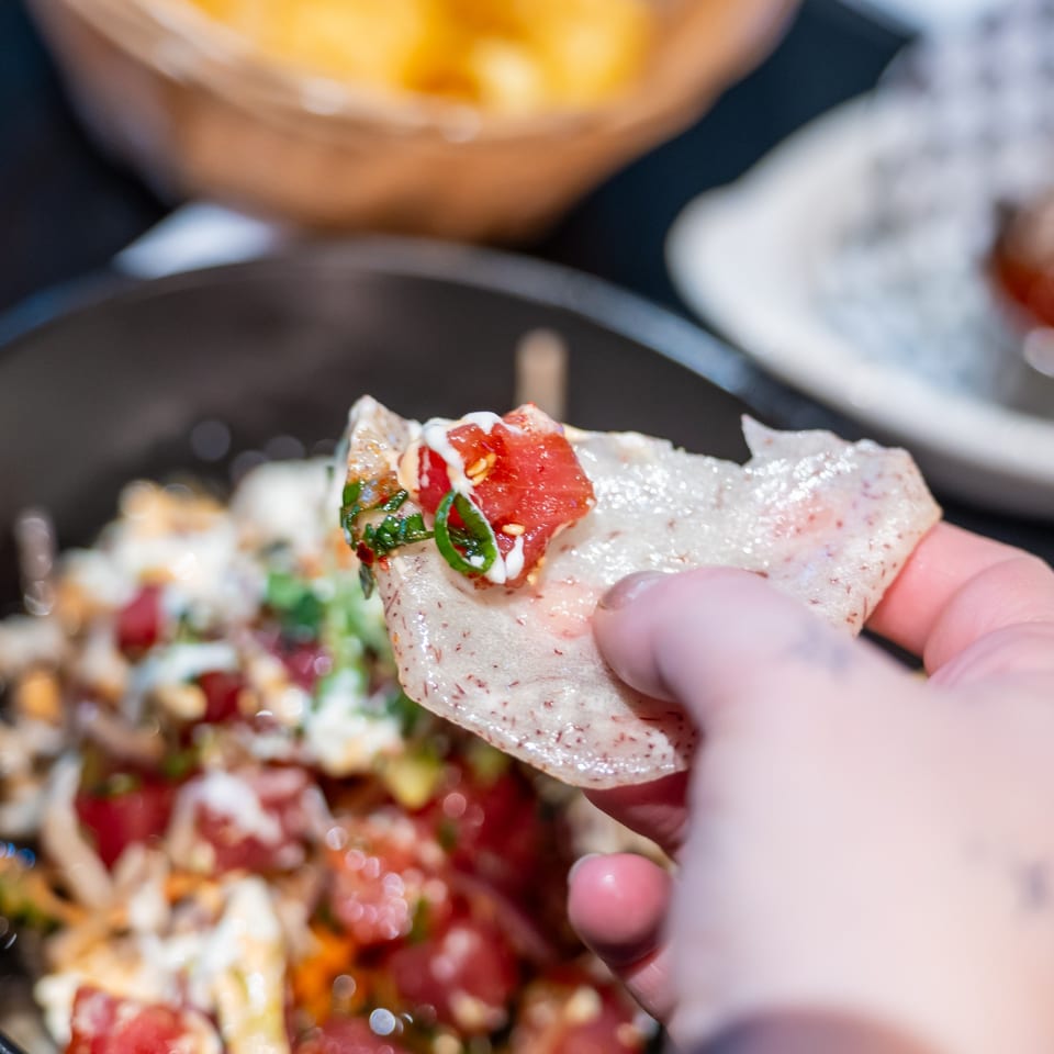 Hand holding a chip with tuna poke, white sauce, and green onions over a blurred bowl.